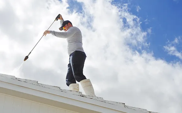 A professional cleaner using a long-handled pressure washer to clean a white roof under a clear blue sky.
